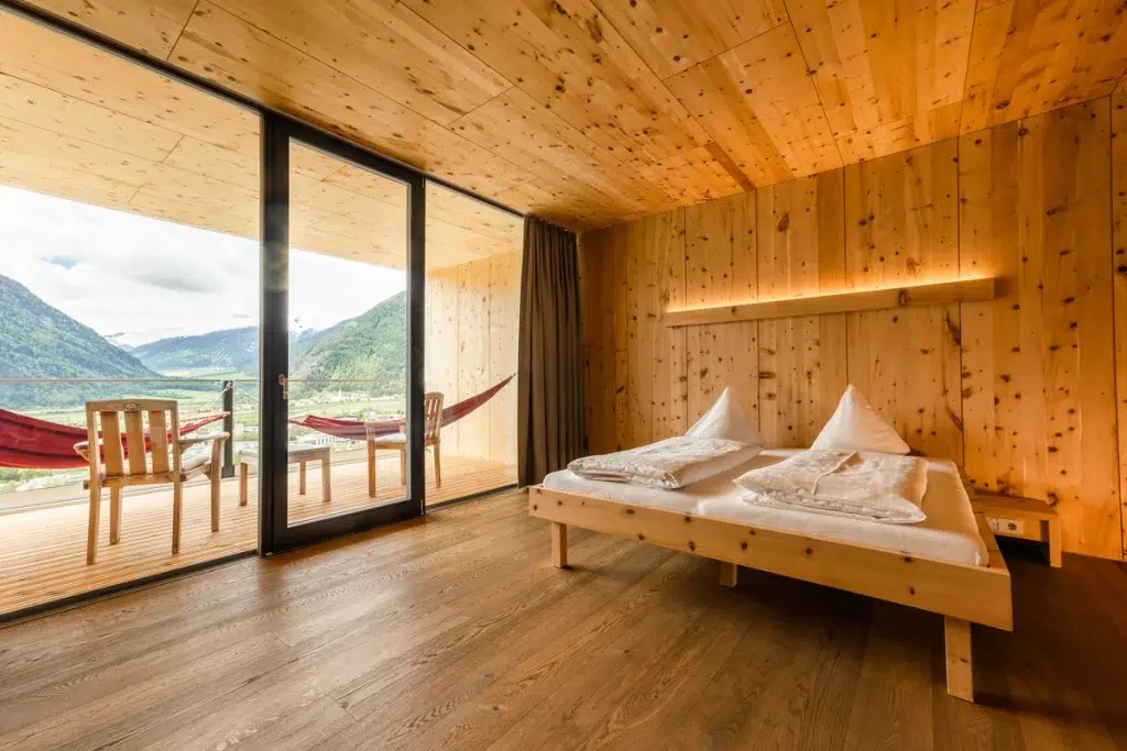 Schlafzimmer aus Holz mit großen Glastüren, die auf einen Balkon mit Hängematten und Bergblick führen.