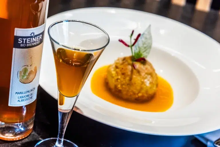 Ein Glas bernsteinfarbener Likör neben einem Dessert mit Soße und Garnitur auf einem weißen Teller.