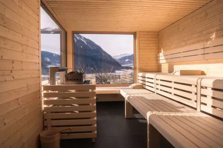 Holzsauna mit Bänken, großes Fenster mit Blick auf verschneite Berge und einen hellen, sonnigen Himmel.
