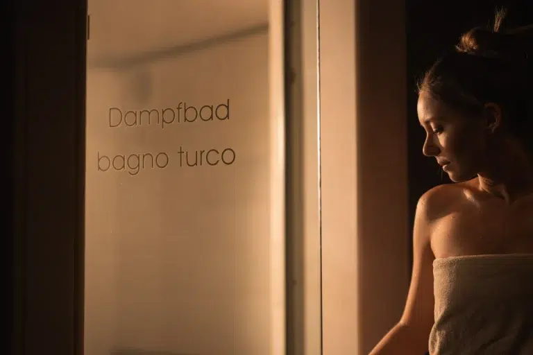Eine in ein Handtuch gewickelte Frau steht vor einer Glastür mit der Aufschrift Dampfbad bagno turco.