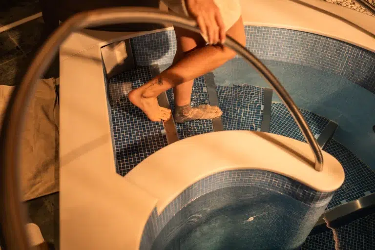 Eine Person in einem Handtuch steigt in ein gekacheltes Hallenbad und hält sich an einem Metallgeländer fest.