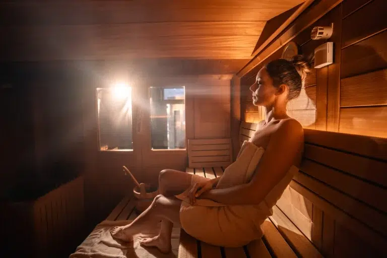 Eine Frau entspannt sich in einer Holzsauna, eingewickelt in ein Handtuch, während das Sonnenlicht hereinstrahlt.