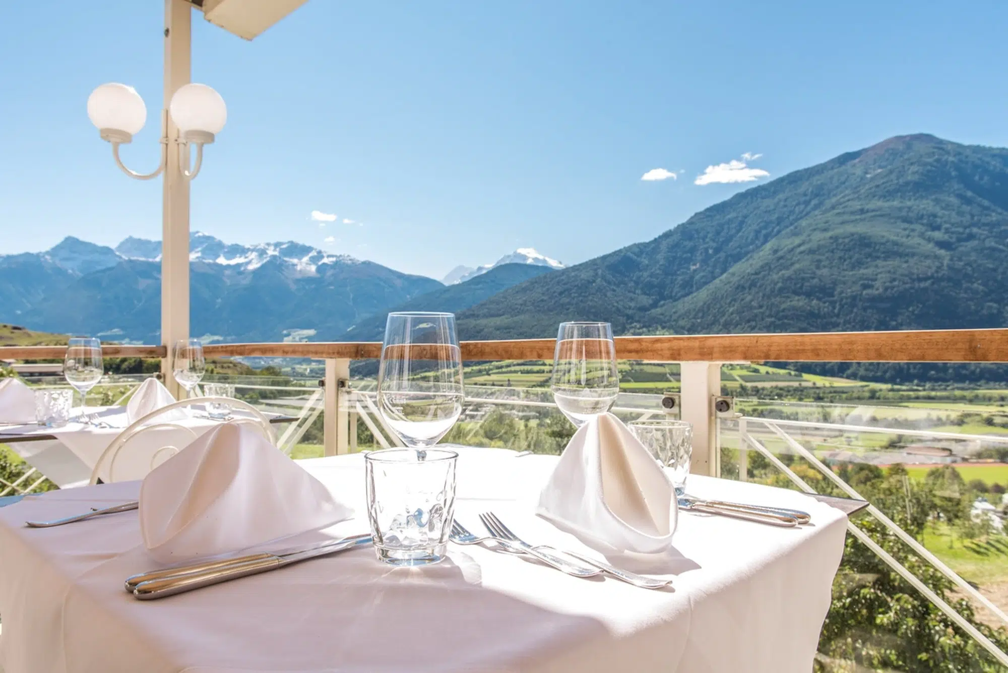 Eleganter Esstisch im Freien mit Glasgeschirr und Blick auf die Berge und das grüne Tal.