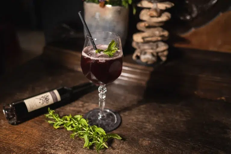 Ein dunkler Cocktail in einem Weinglas mit Minzgarnitur, daneben eine Weinflasche und frische Minze.