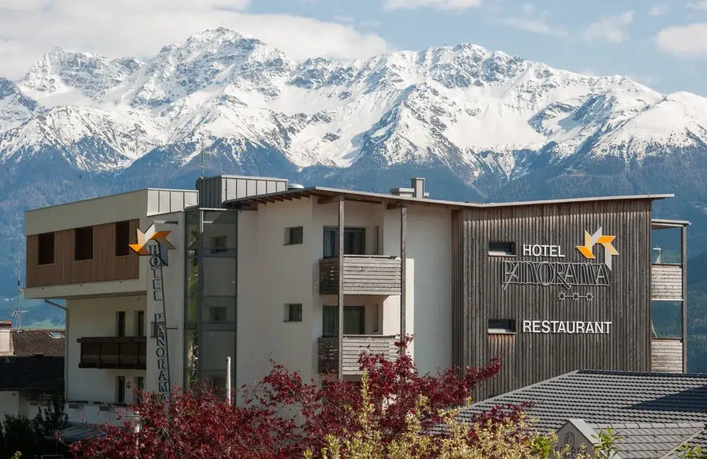 Modernes Hotelgebäude mit Balkonen vor schneebedeckten Berggipfeln im Hintergrund.