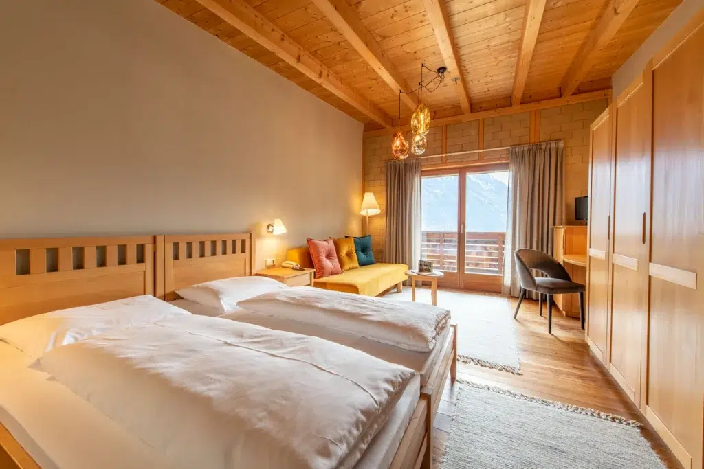 Gemütliches Hotelzimmer mit zwei Betten, Holzmöbeln und Bergblick durch Glastüren.