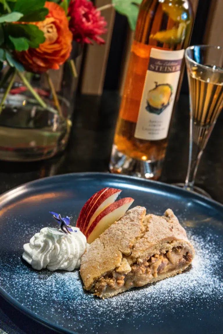 Scheibe Apfelstrudel mit Schlagsahne und Apfelspalten, mit Wein und Blumen im Hintergrund.