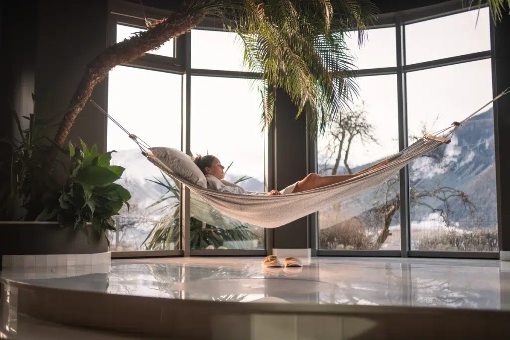 Eine Person entspannt sich in einer Hängematte im Haus, in der Nähe großer Fenster mit Blick auf die verschneiten Berge.