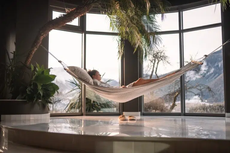 Eine Person entspannt sich in einer Hängematte im Haus, in der Nähe großer Fenster mit Blick auf die verschneiten Berge.