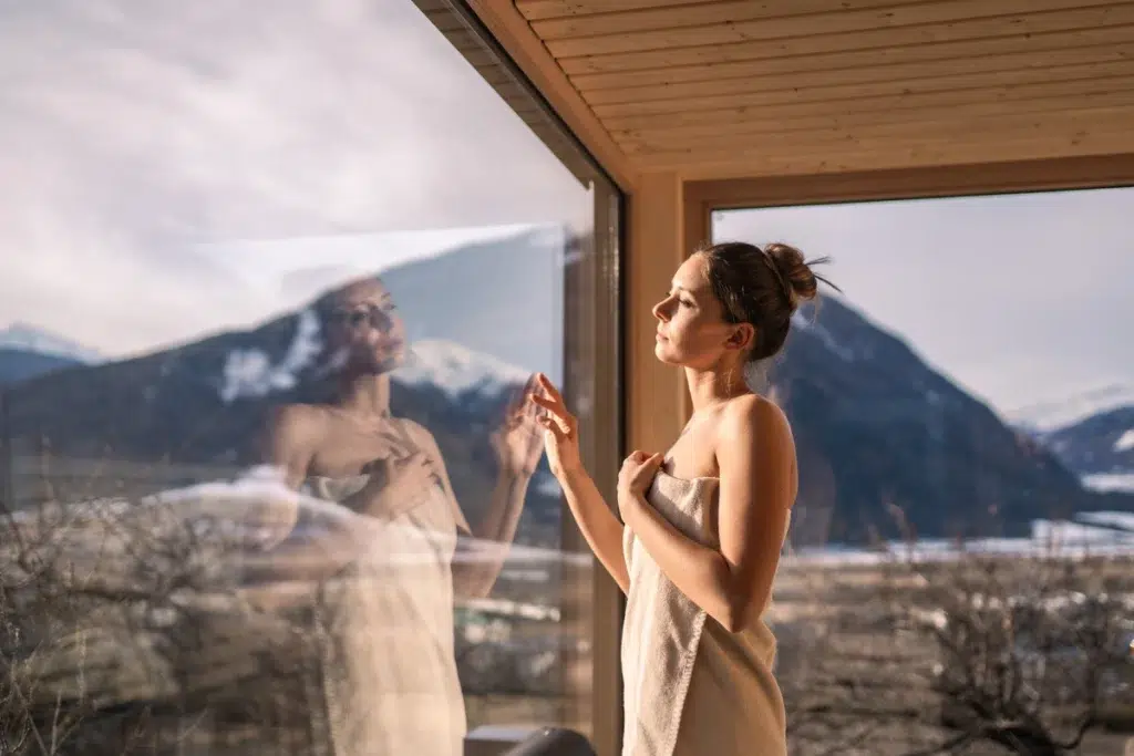 Frau in einem Handtuch im Biohotel Südtirol, Berge und ihr Spiegelbild am Fenster sichtbar.