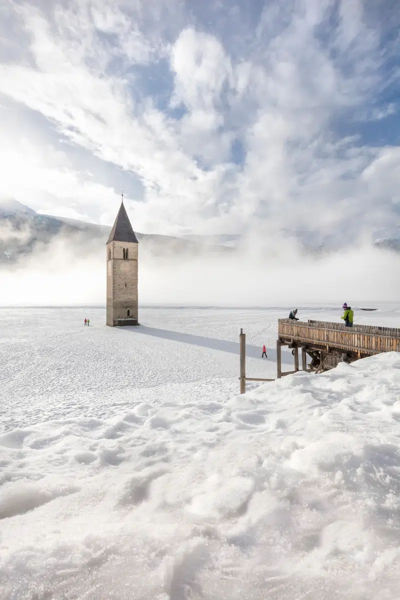 Ein Kirchturm erhebt sich aus einem zugefrorenen, schneebedeckten See mit Menschen in der Nähe und bewölktem Himmel.