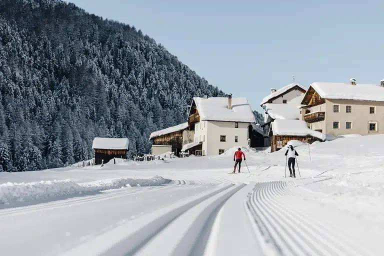 Zwei Personen beim Langlaufen in der Nähe von Almhütten, einem bewaldeten Hügel und einem Biohotel Südtirol.