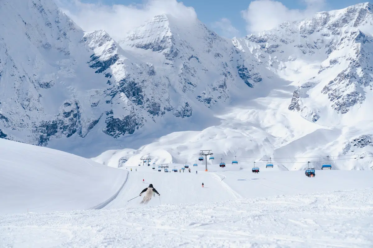 Ein Skifahrer fährt einen verschneiten Berghang hinunter, mit Sesselliften und Gipfeln im Hintergrund.