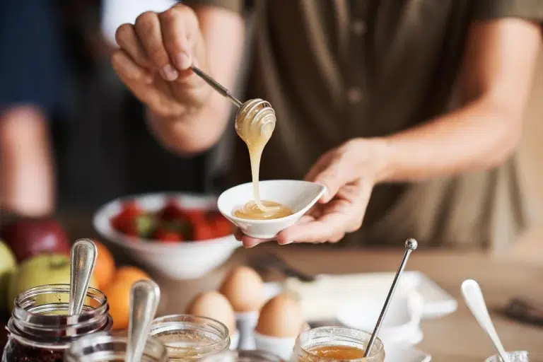 Eine Person träufelt Honig in eine kleine Schale mit Gläsern, Eiern und Früchten im Hintergrund.
