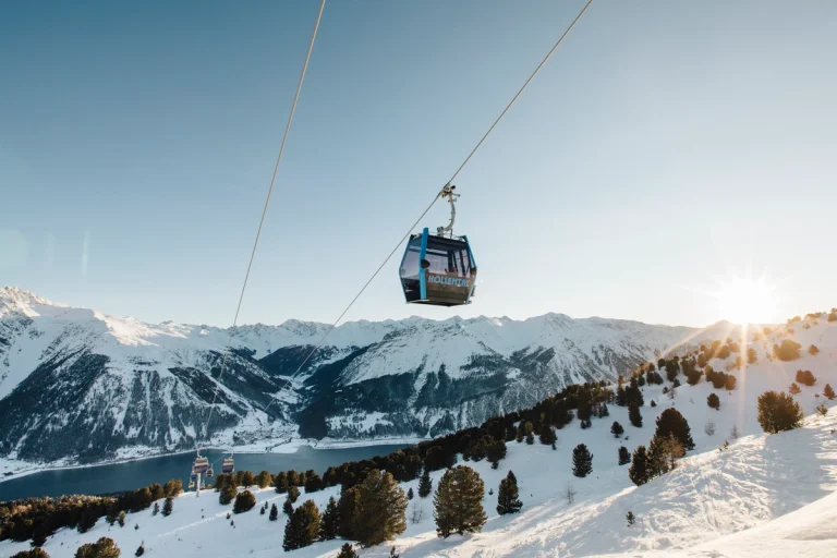 Eine Seilbahn fährt bei Sonnenaufgang über schneebedeckte Berge, unter denen sich Pinienbäume befinden.