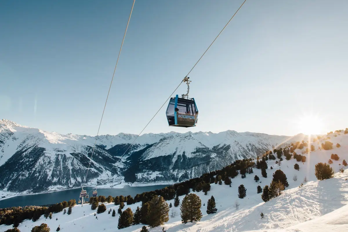 Eine Seilbahn fährt bei Sonnenaufgang über schneebedeckte Berge, unter denen sich Pinienbäume befinden.