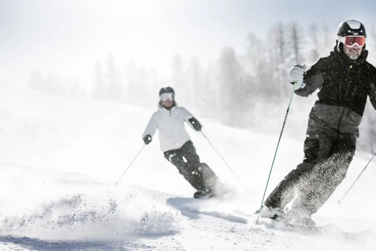 Zwei Personen fahren auf einer verschneiten Piste mit Bäumen im Hintergrund bergab.