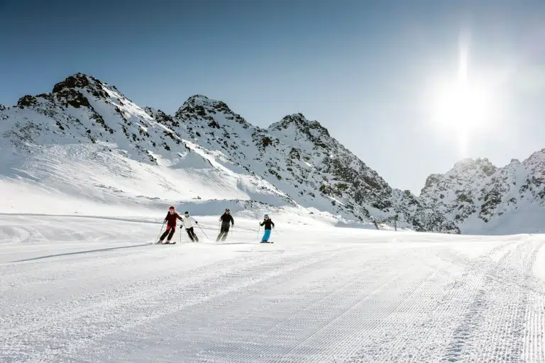 Drei Skifahrer auf einer verschneiten Piste mit Bergen und heller Sonne im Hintergrund.