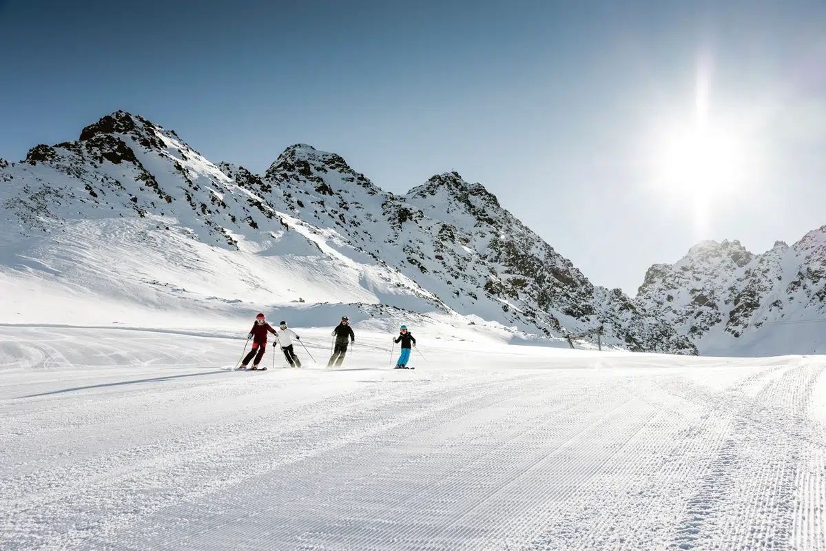 Drei Skifahrer auf einer verschneiten Piste mit Bergen und heller Sonne im Hintergrund.