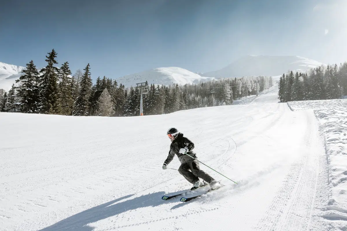 Person beim Skifahren auf einer verschneiten Piste mit Bäumen und Bergen im Hintergrund.