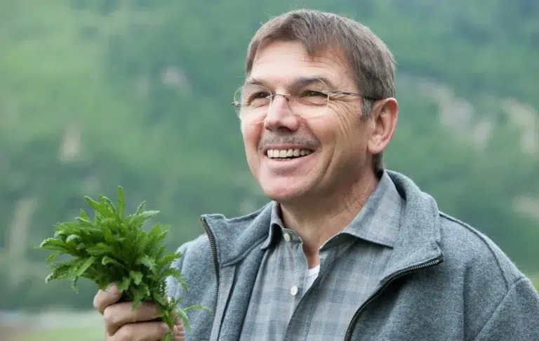 Lächelnder Mann, der einen Strauß frischer grüner Kräuter im Freien hält, mit Bergen im Hintergrund.