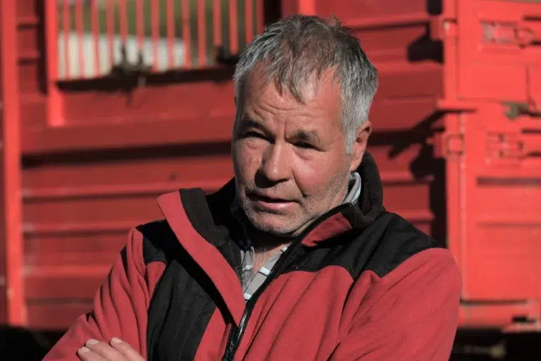 Karl Thöni Mals, älterer Mann in rot-schwarzer Jacke steht draußen bei einem roten Bauwerk.