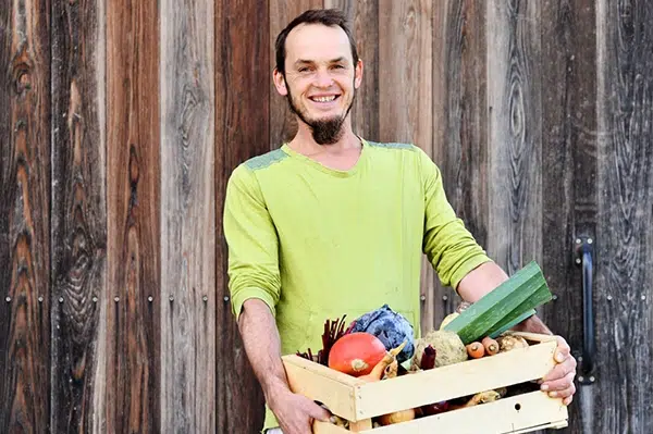 Daniel Primisser hält lächelnd eine Kiste mit verschiedenen Gemüsesorten vor eine Holzwand.