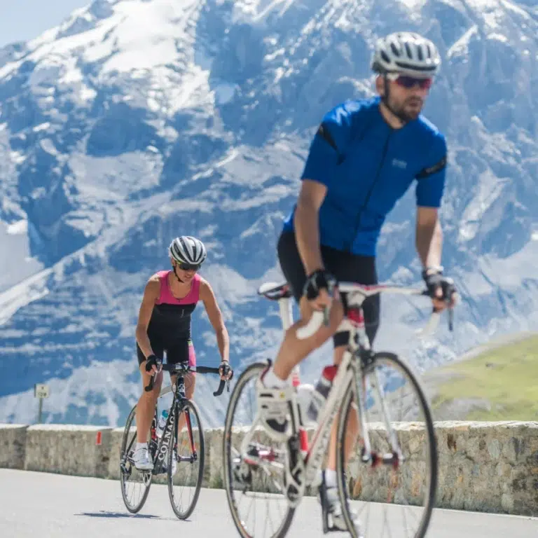 Zwei Radfahrer fahren auf einer Bergstraße im Vinschgau mit verschneiten Gipfeln im Hintergrund.