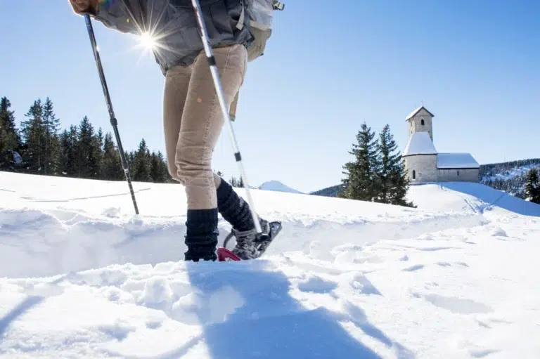 Person beim Schneeschuhwandern im hellen Schnee in der Nähe einer kleinen Kirche und von Tannenbäumen in Langlaufen im Vinschgau.