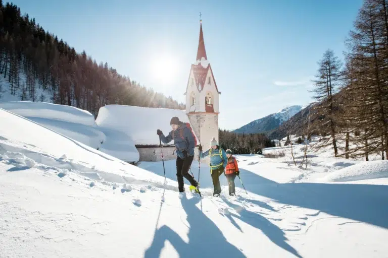 Drei Personen wandern im Tiefschnee in der Nähe einer Kirche und genießen die Landschaft des Vinschgaus von Langlaufen.