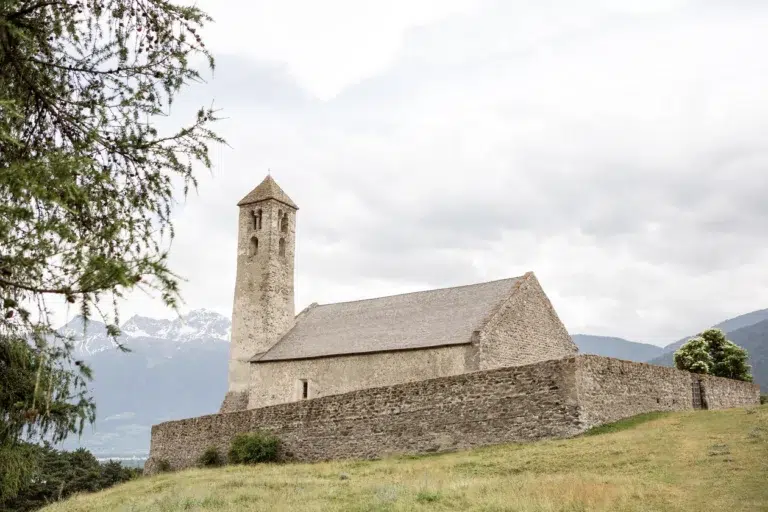 Kleine Steinkirche mit einem Turm, umgeben von einer Steinmauer, auf einem grasbewachsenen Hügel gelegen.