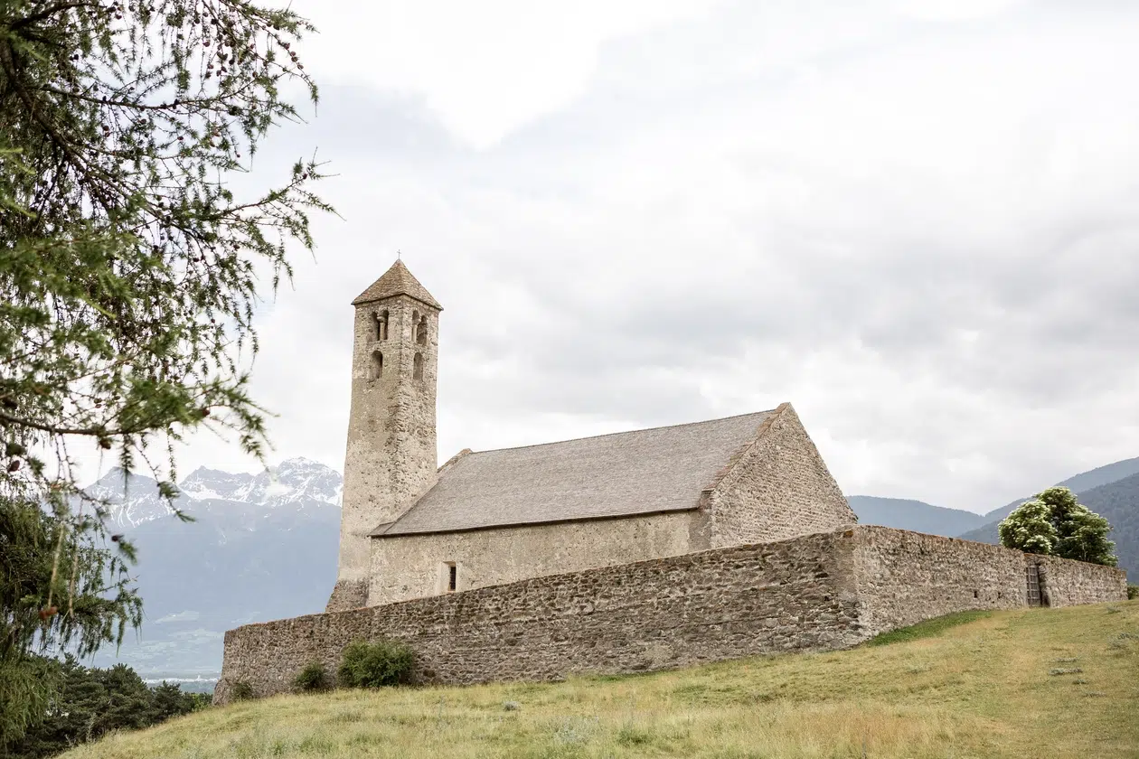 Kleine Steinkirche mit einem Turm, umgeben von einer Steinmauer, auf einem grasbewachsenen Hügel gelegen.