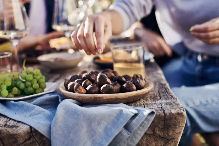Eine Hand greift nach Kastanien auf einem Holzteller an einem Tisch im Freien mit Getränken und Weintrauben.