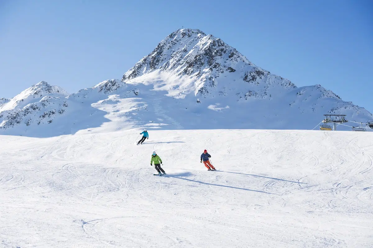 Drei Skifahrer fahren bei strahlend blauem Himmel einen verschneiten Berghang hinunter.