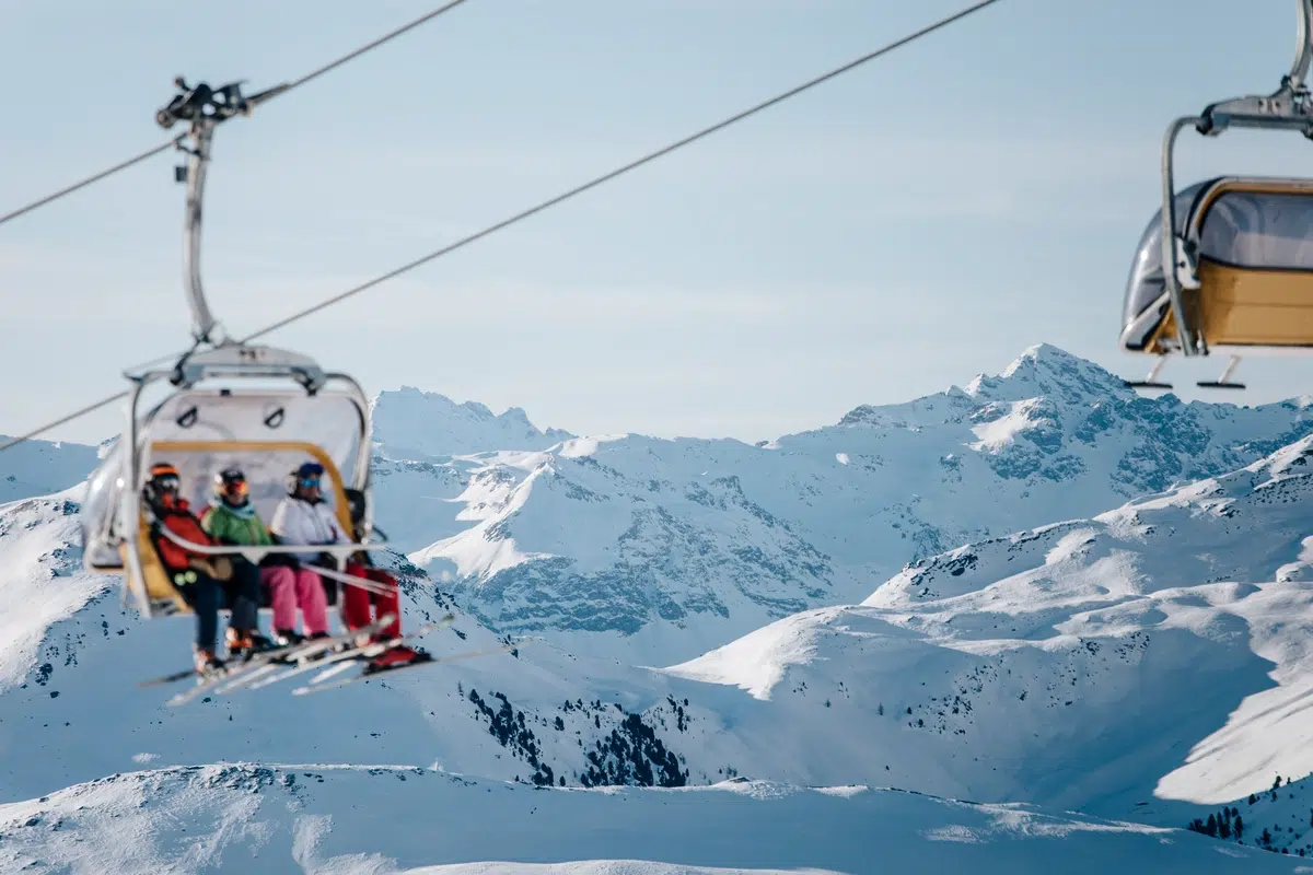 Skifahrer in einem Sessellift mit verschneiten Bergen im Hintergrund.