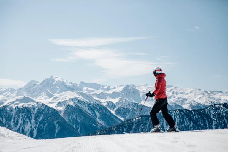 Skifahrer in einer roten Jacke steht auf einer verschneiten Piste mit Bergen im Hintergrund unter klarem Himmel.