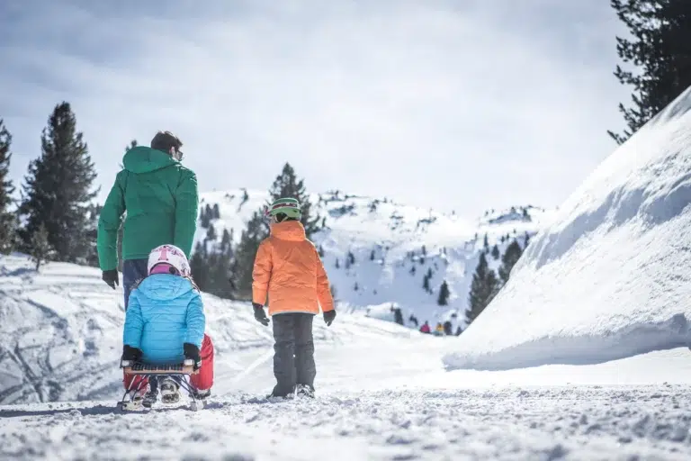 Ein Erwachsener und zwei Kinder rodeln in der Nähe von Langlaufen Vinschgau in einer verschneiten Berglandschaft.