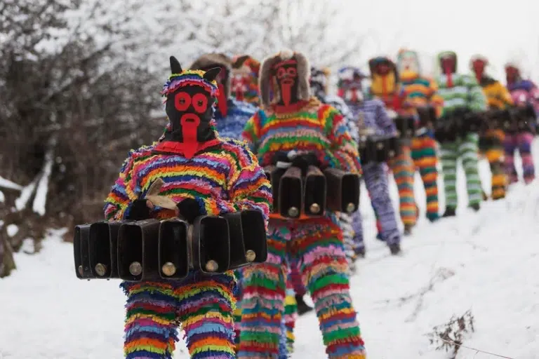 Menschen in bunten, gestreiften Kostümen und Masken laufen durch den Schnee und tragen große Glocken.