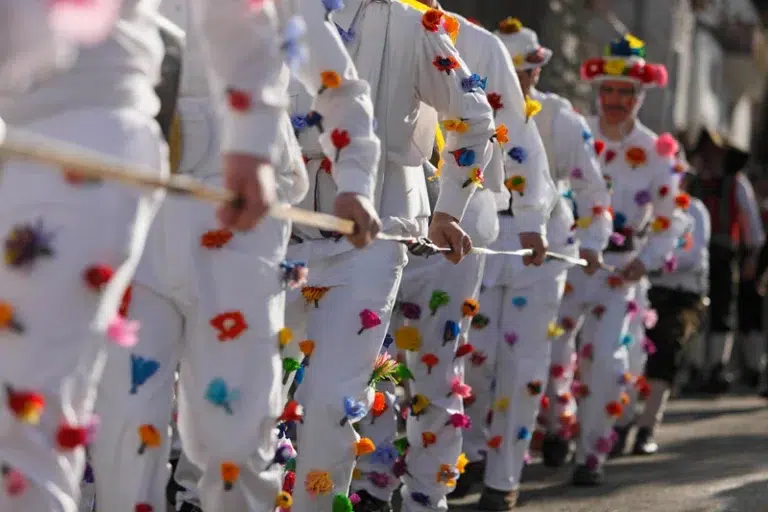 Menschen in weißen Kostümen mit bunten Bommeln gehen in einer Reihe und halten einen langen Stock.
