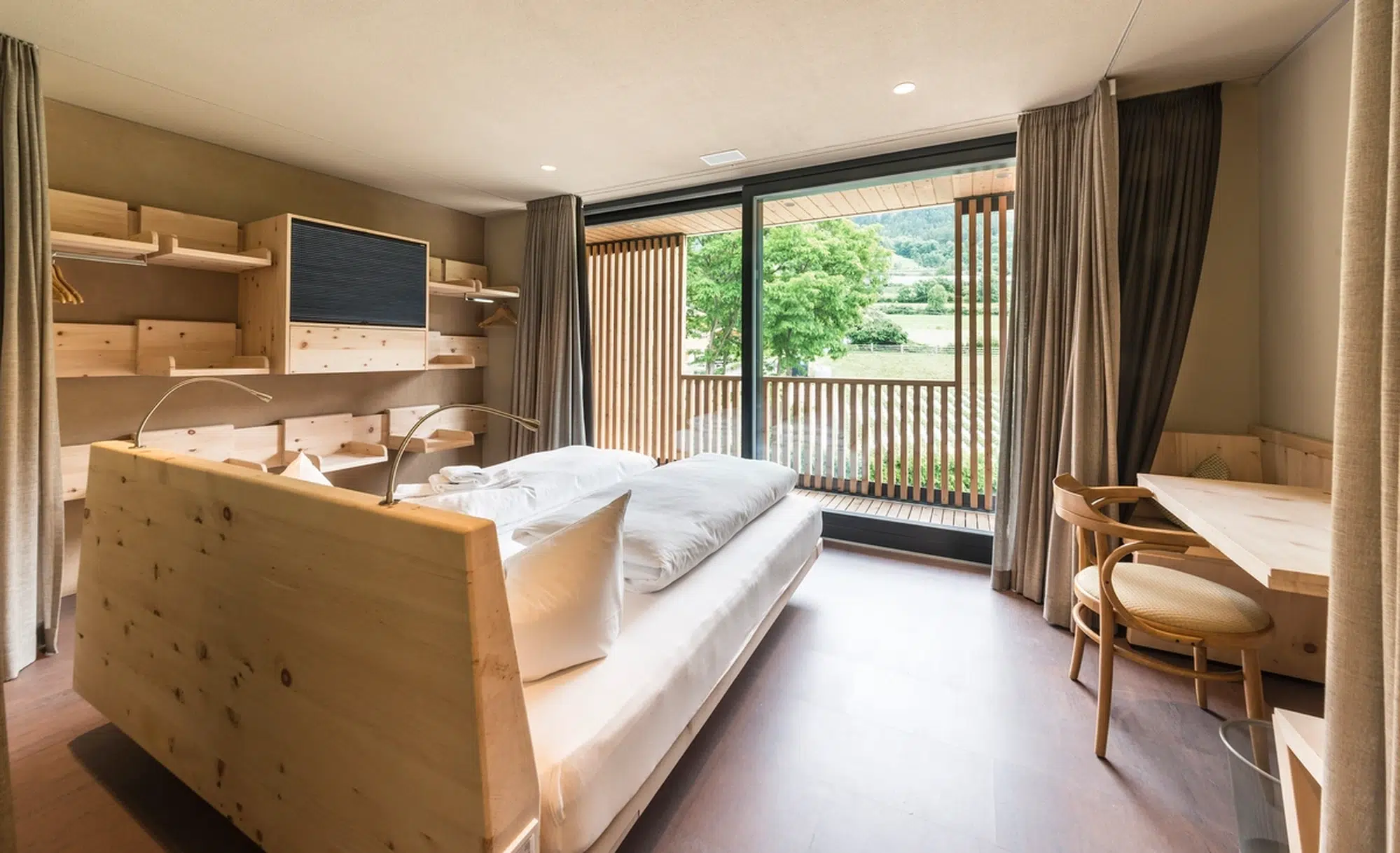 Modernes Hotelzimmer mit Holzmöbeln, großem Bett, Schreibtisch und Balkon mit Blick ins Grüne.