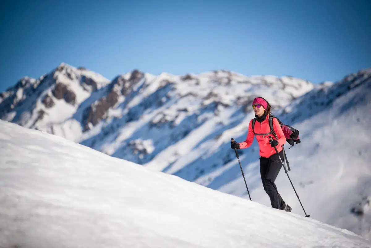 Persona in tenuta invernale che fa escursioni in salita su una montagna innevata con bastoncini; sullo sfondo cime innevate.