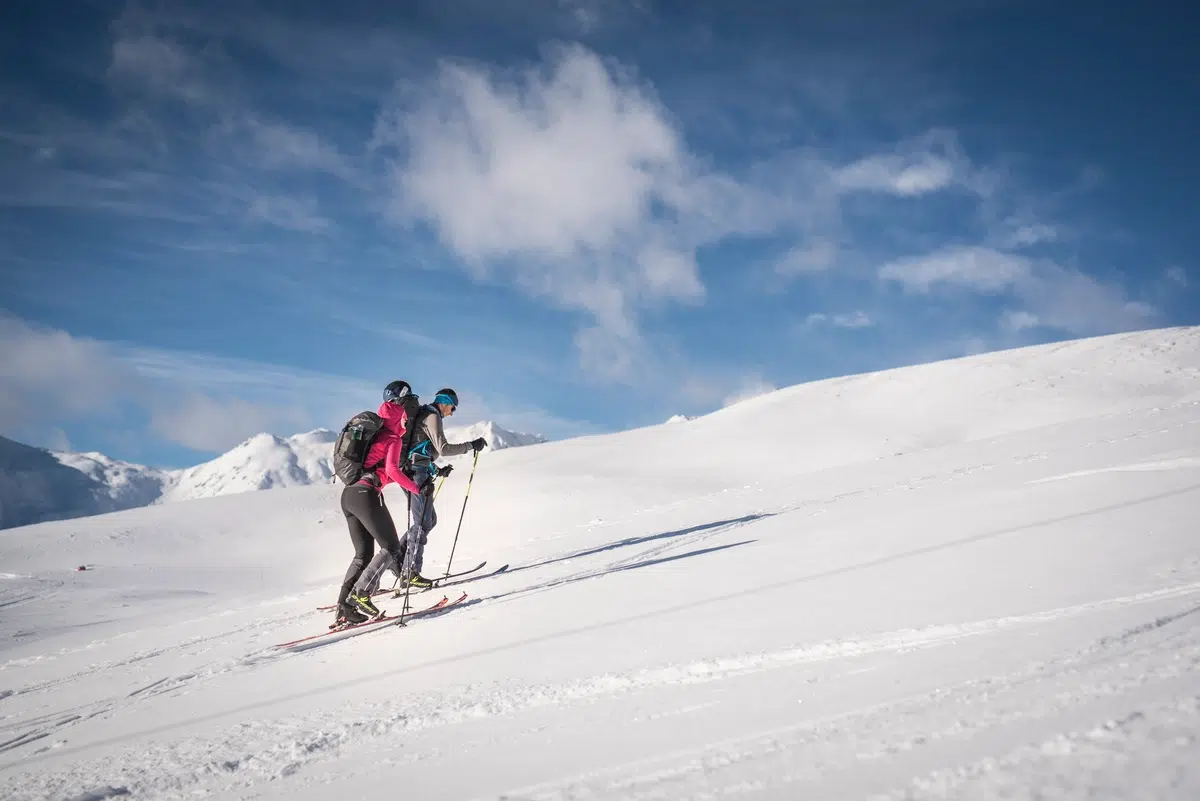Due persone che sciano in salita su una montagna innevata sotto un cielo blu con nuvole sparse.