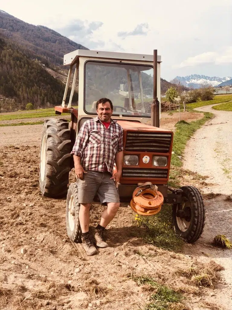 Mann im karierten Hemd, der neben einem alten roten Traktor auf einem Feldweg mit Bergen im Hintergrund steht.