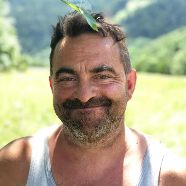 Lächelnder Mann im Freien mit einem Blatt auf dem Kopf, in einem Tank-Top, mit Grünzeug hinter ihm.