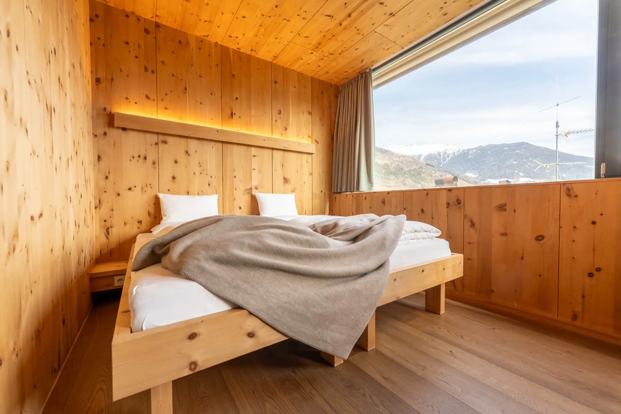 Gemütliches Holzzimmer mit großem Fenster, Blick auf die Berge und einem ungemachten Bett mit Decken.