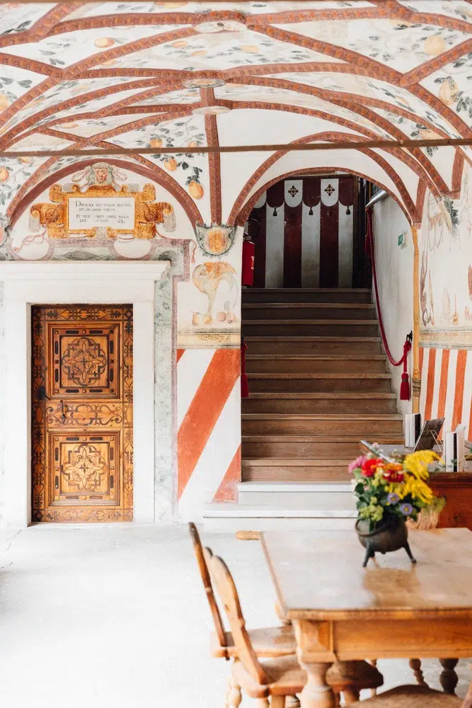 Corridoio decorato con soffitto dipinto, porta in legno intagliato e scale accanto a un tavolo con fiori.