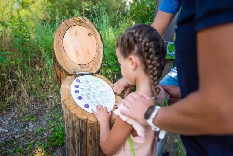 Una bambina legge un cartello su un palo di legno all'aperto, guidata dalla mano di un adulto sulla sua spalla.