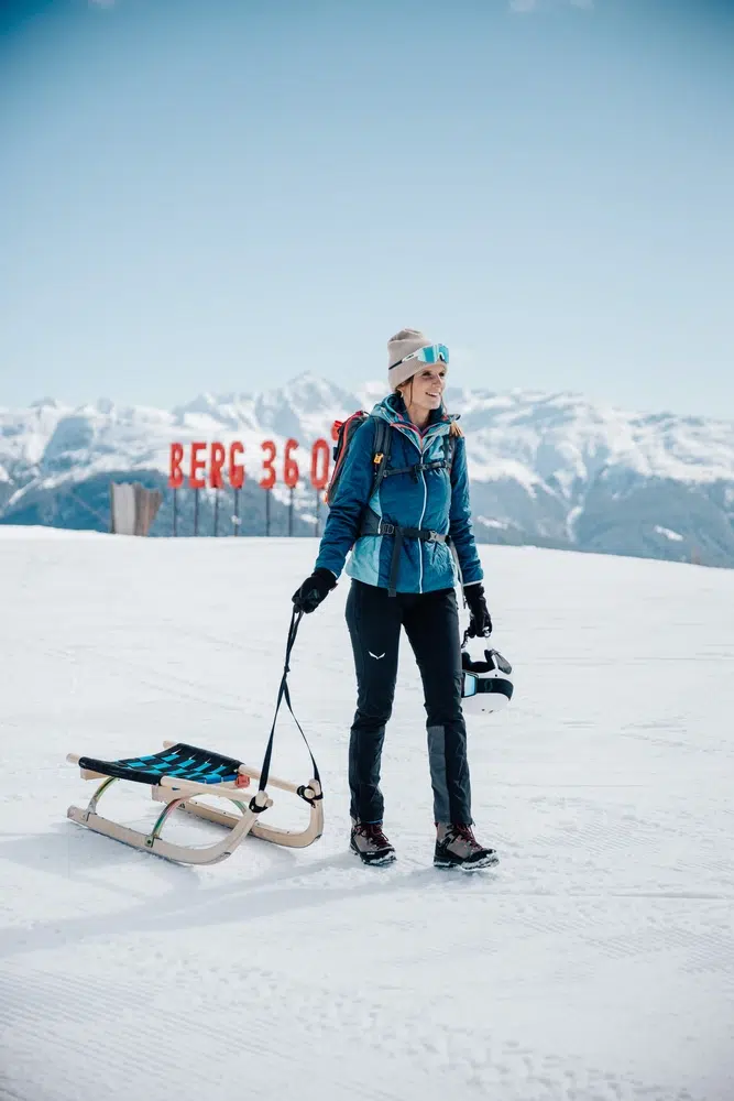 Persona in tenuta invernale in piedi su una montagna innevata, con slitta in mano e montagne sullo sfondo.