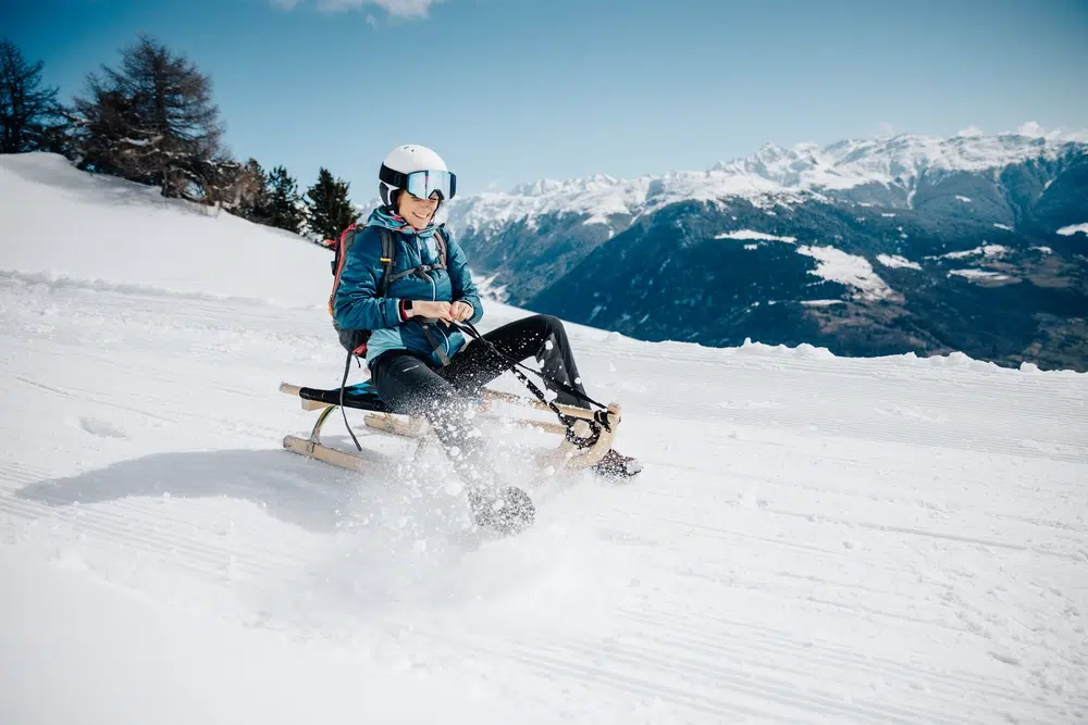 Persona che scende con lo slittino da un pendio innevato con le montagne sullo sfondo.