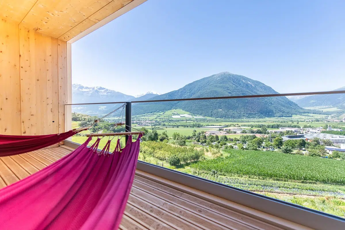 A red hammock on a wooden balcony overlooks green fields and a mountain under blue sky.